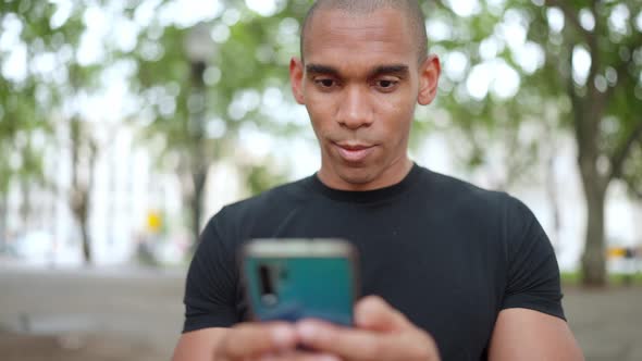 Positive bald African wearing black t-shirt typing on phone while sitting on the bench