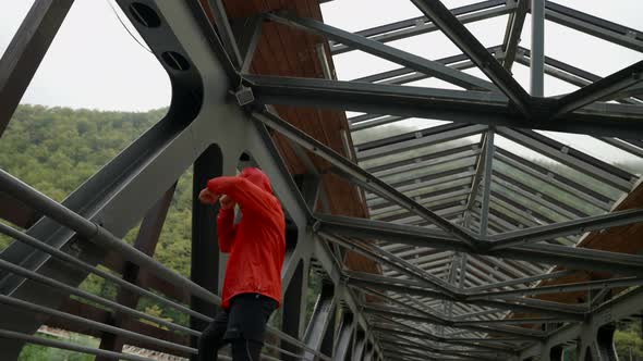 a Man in a Red Jacket with a Hood is Boxing on a Railway Bridge