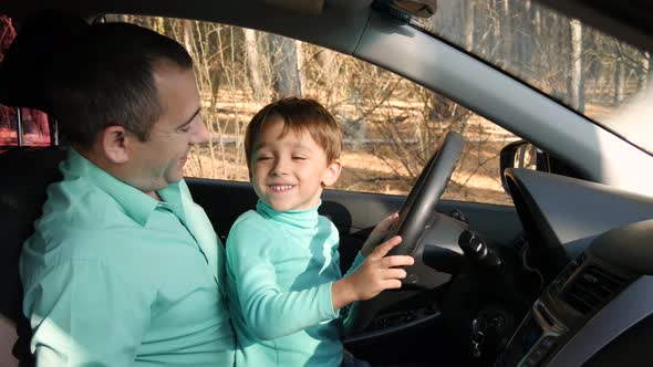 A Cute Boy Turns the Wheel of a Car, Sitting on His Father's Lap. The Father Teaches His Son the