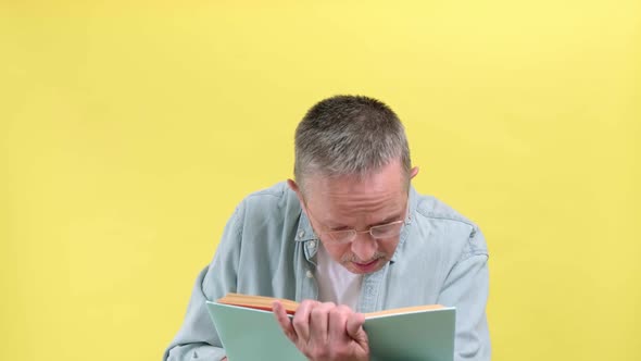 Aged Man with Blue Shirt Holds Open Book in Her Hands on a Yellow Background