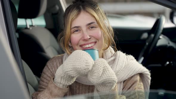 Happy young caucasian woman with thermo cup sitting inside the car in winter and warming up