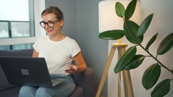 Woman with Glasses Sits on a Sofa in a Cozy Room and Makes an Online Shopping Using a Credit Card