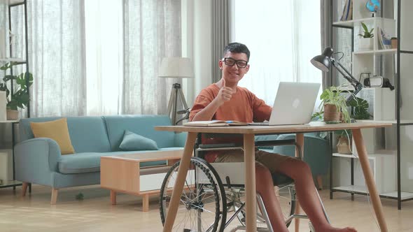 Asian Boy Sitting In A Wheelchair Using Laptop Computer While Smiling And Thumb Up To Camera At Home