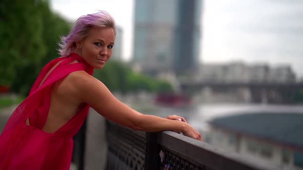 Pink Haired Woman in Red Dress Is Walking on City Embankment in Summertime at Windy Weather