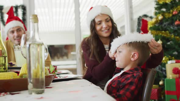 Happy caucasian mother putting on santa hat to son at christmas table
