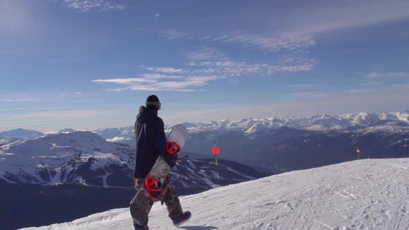 A young man snowboarder walking with his board on a scenic snow covered mountain top.