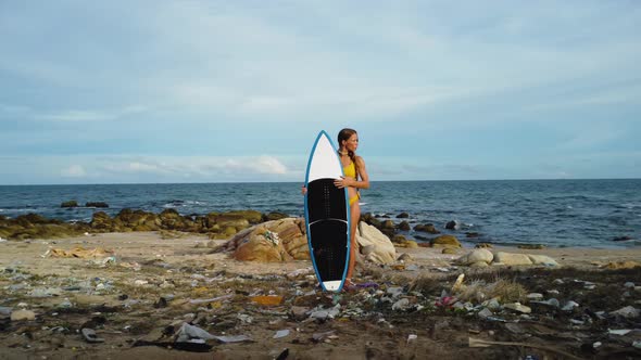 Young sexy female surfer in bikini holding surfboard on polluted ocean beach filled with plastic was