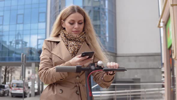 Young Stylish Blonde Woman in a Beige Trench Coat and Jeans Takes Electric Kick Scooter in Sharing