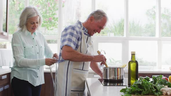 Senior woman putting apron on her husband while he cooks in the kitchen at home