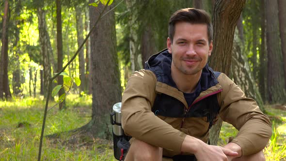 A Young Hiker Sits on the Ground in Forest and Shows a Thumb Up To the Camera with a Smile
