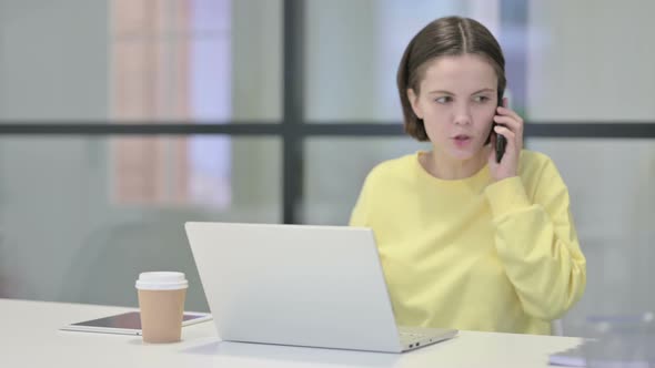 Angry Woman Talking on Smartphone While Using Laptop in Office