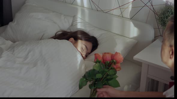 Young Female Woman Sleeping in Bed and Loving Man Putting Bunch Roses on Sheet