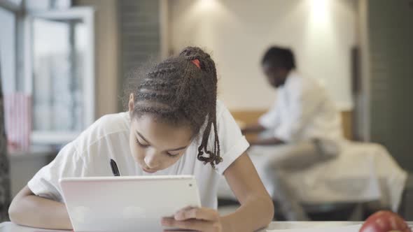 Concentrated African American Teenage Girl Doing Homework Home Blurred Man Sitting Bed Background