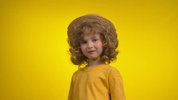 Cute Little Curly Girl in a Straw Hat Smiles Thoughtfully in a Yellow Background