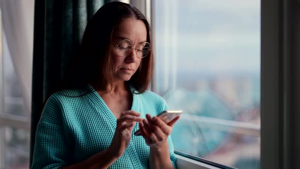 A Businesslike Adult Woman with Glasses Standing at the Window of the House in the Morning in a