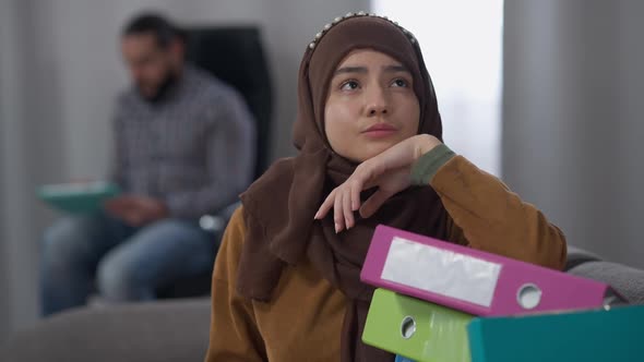 Sad Tired Young Woman in Hijab Sighing Thinking Indoors with Blurred Middle Eastern Man Surfing