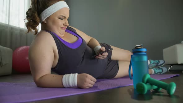 Fat Lazy Woman Lying on Floor With Smartphone in Hands Instead of Training