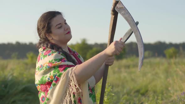 Portrait of Beautiful Overweight Woman Sharpening Scythe Preparing To Start Mow on the Green Summer