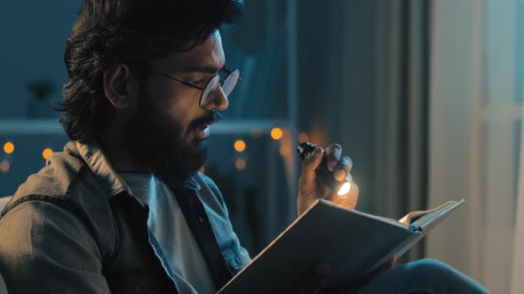 Bearded Adult Man in Glasses at Night Home Living Room with Flashlight Shines on Book Reads