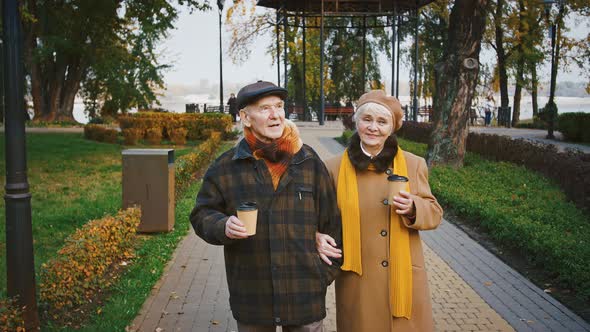 Happy Pair Grandparents Holding Paper Cups of Coffee Talking and Smiling During a Walk in Autumn