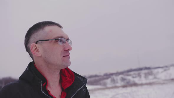 An Adult Man with Glasses Looks Thoughtfully Into the Distance Standing on the Street in Winter in