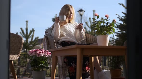 charming woman with phone drinking tea on the terrace with flowers