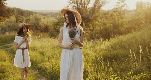 Two Girl Teenagers in White Dresses and Straw Hats with Flowers Bouquet Posing on Nature Background