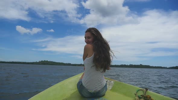 Beautiful Smiling Girl with Blowing Brown Hair Sits on Bow of Boat and Looks Into Camera. Happy