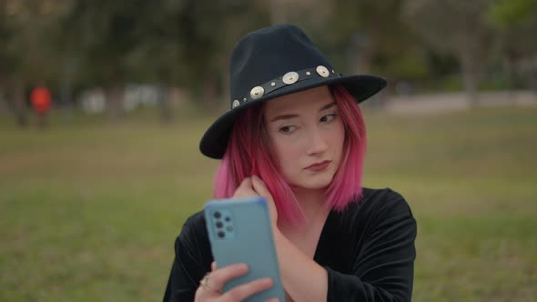 Pleasantlooking Caucasian Girl with Pink Hair and Black Hat Looks at Herself on Cell Phone