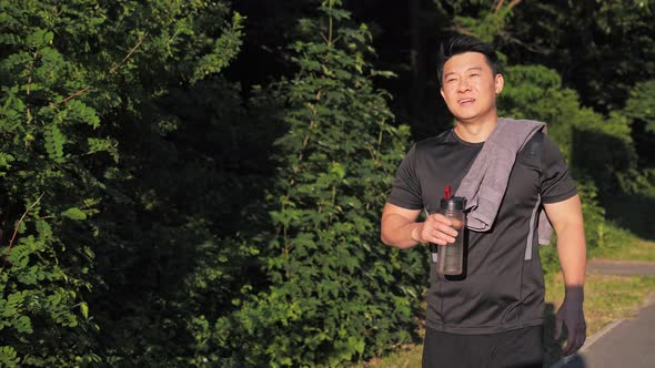 Happy Asian Man with Towel Walking in Summer Park Drinking Water After Running