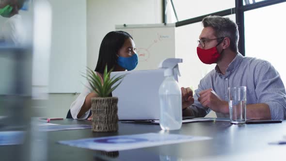 Diverse male and female office colleagues wearing face masks discussing over laptop at modern office