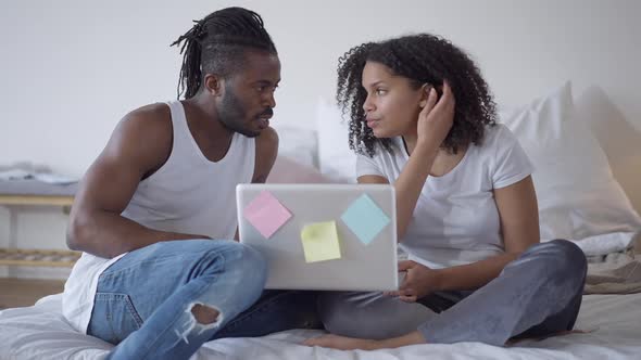 Wide Shot Portrait of Concentrated African American Young Couple Sitting on Bed Discussing Business