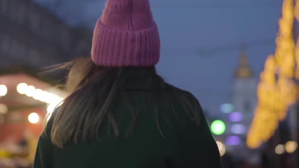 Camera Following Young Caucasian Woman in Pink Hat and Green Coat Walking To Children's Carousels