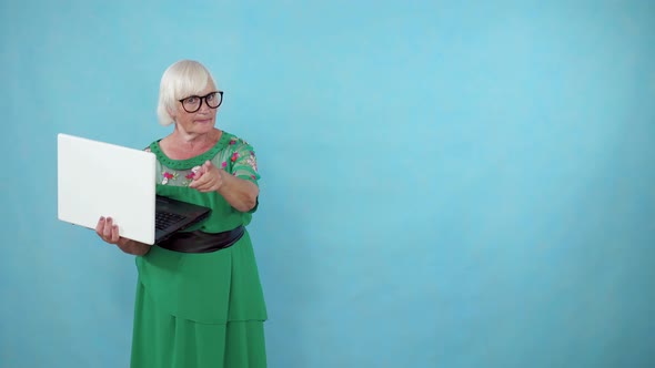 Thoughtful Old Woman with a Laptop in Her Hands Finds a Solution and Stands on a Blue Background