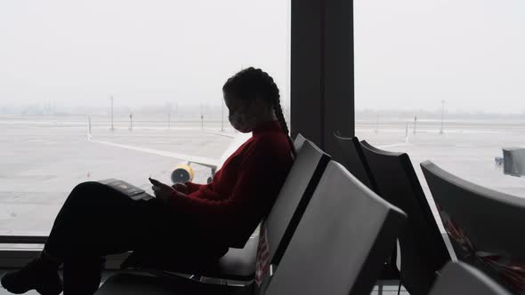 Masked Young Woman in Airport Waiting Room Sits and Using a Smartphone By Window