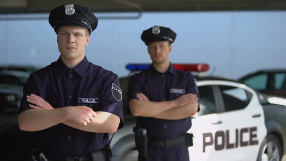 Serious Male Officers Standing With Hands Crossed Against Police Car Background