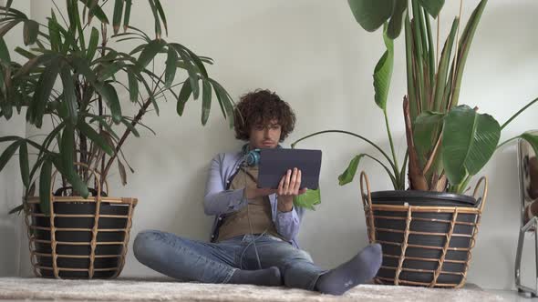 Relaxed Young Man with Curly Hair Browsing the Internet on His Tablet Computer As He Sits on on the