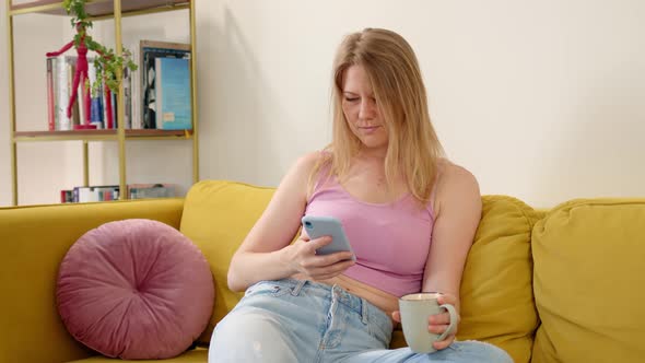 Nice Woman in Pink Tank Top and Blue Jeans Sits on Yellow Sofa and Drinks Hot Tea or Coffee From