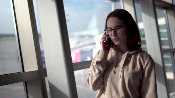 A Young Woman in a Sweatshirt and Glasses Walks Through the Airport and Speaks on the Phone Against