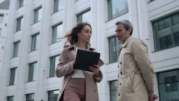A beautiful girl with a tablet approaches the head of a man standing near office buildings