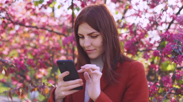 Attractive Young Redhaired Woman Uses Smatphone in Park at Sunset