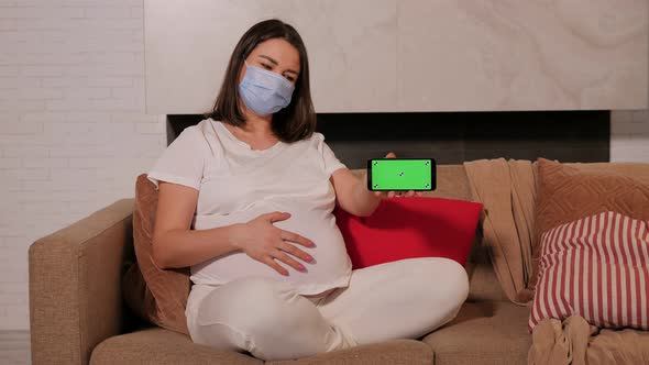 Pregnant Woman in a Mask on the Sofa She Shows a Phone with a Green Screen