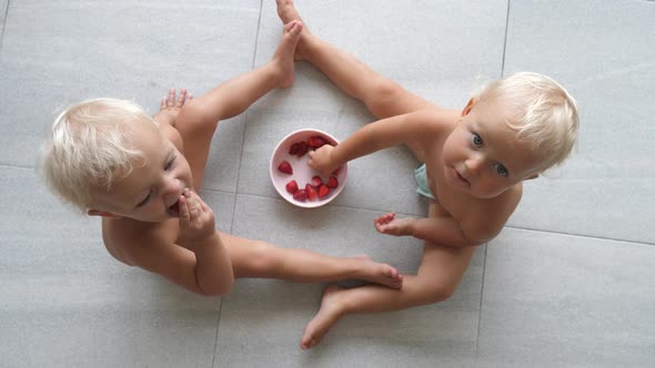 Top View of Cute Twins Sitting on the Floor Eating Strawberries From Shared Cup