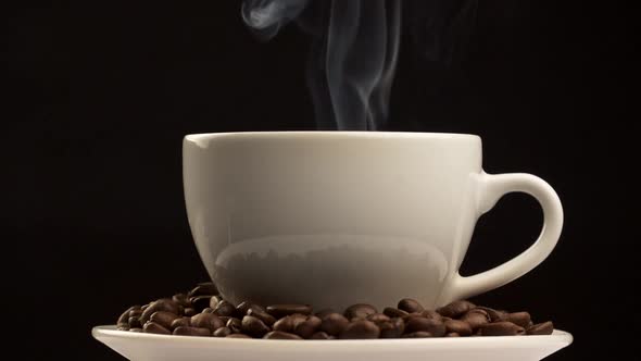 Steam Coming Out of Cup of Coffee on a Saucer Filled with Brown Fried Coffee Beans. Black Background