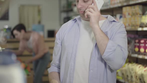 Close-up of Bored Caucasian Husband Talking on the Phone with Wife As Choosing Products in Grocery