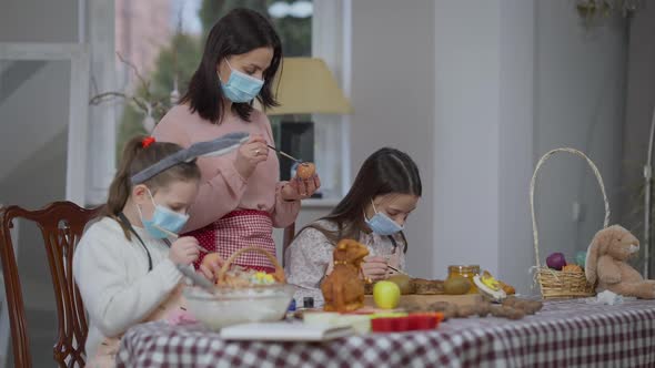 Focused Caucasian Woman and Girls in Coronavirus Face Masks Coloring Eggs on Easter Indoors