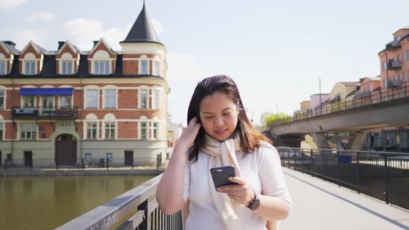 Asian black straight hair woman walking on a bridge over a river in town and using her smartphone