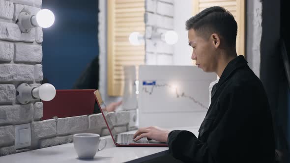 Focused Asian Man Typing on Laptop Keyboard Analyzing Graph in Home Office at Night