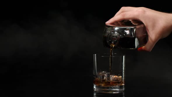 Barman Pour Whiskey Cognac Brandy From Bottle Into Drinking Glass with Ice Cubes on Dark Background