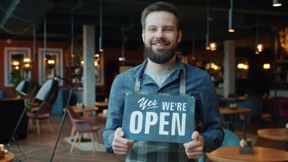 Portrait of Cheerful Handsome Young Waiter in Apron Holding We Are Open Sign in Coffee Shop